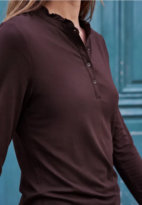 Eine Frau trägt ein braunes Langarm-Henleyshirt mit Knopfleiste vor einer türkisfarbenen Wand.