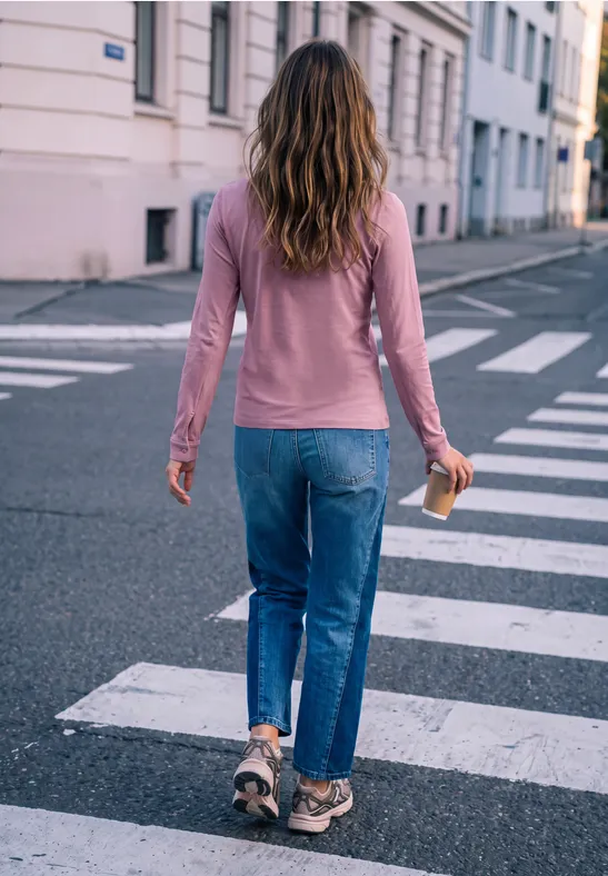 Eine Frau in Jeans und pinkem Pullover überquert mit einem Coffee-to-go-Becher die Straße auf einem Zebrastreifen.