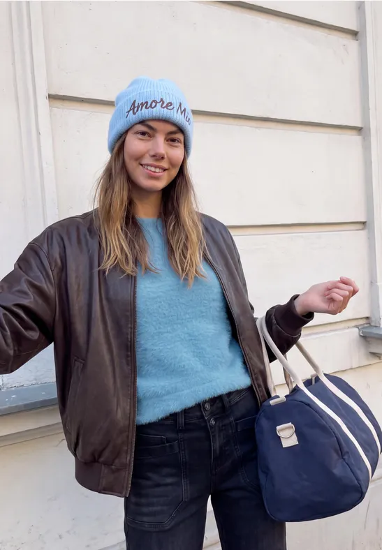 Frau in blauer Strickjacke, Lederjacke und Beanie mit Reisetasche vor heller Wand.