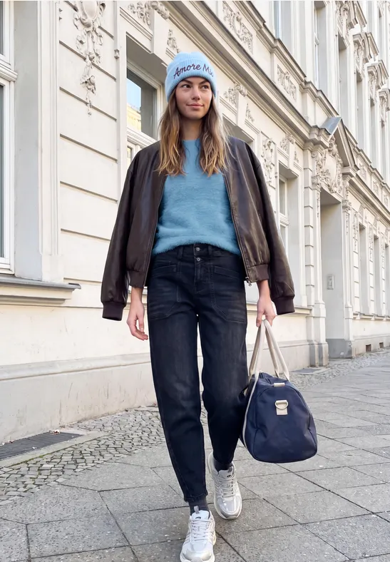 Junge Frau mit blauer Mütze, Pullover und brauner Jacke trägt dunkle Jeans, weiße Sneaker und Tasche auf Bürgersteig.