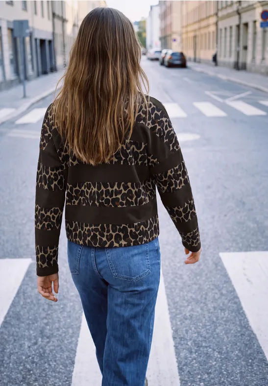 Une personne aux longs cheveux, en jean et pull à motifs, traverse un passage piéton en ville.