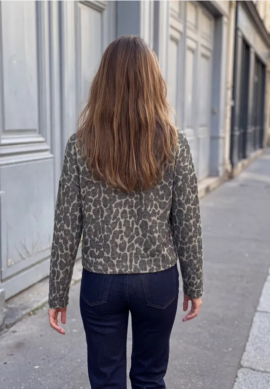 Frau mit langen braunen Haaren, gemusterter Jacke und Jeans geht auf Gehweg neben Häuserfront