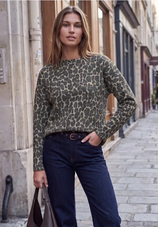 Eine junge Frau in grünem Musterpullover und Jeans posiert mit Tasche auf einer gepflasterten Straßengasse.