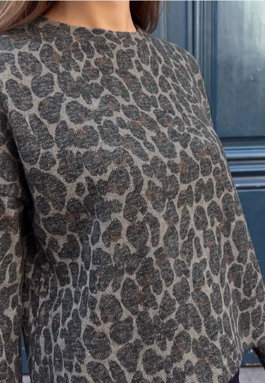 Oberkörper einer Frau in einem grauen Pullover mit Leopardenmuster vor dunklem Hintergrund.