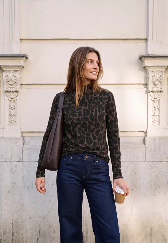 Eine lächelnde Frau mit Kaffeebecher und Schultertasche, in Jeans und Leopard-Top, vor einer Hausfassade.