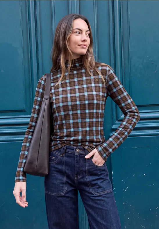 Eine Frau trägt ein kariertes Langarmshirt, Jeans und eine Schultertasche vor einer blauen Holztür.