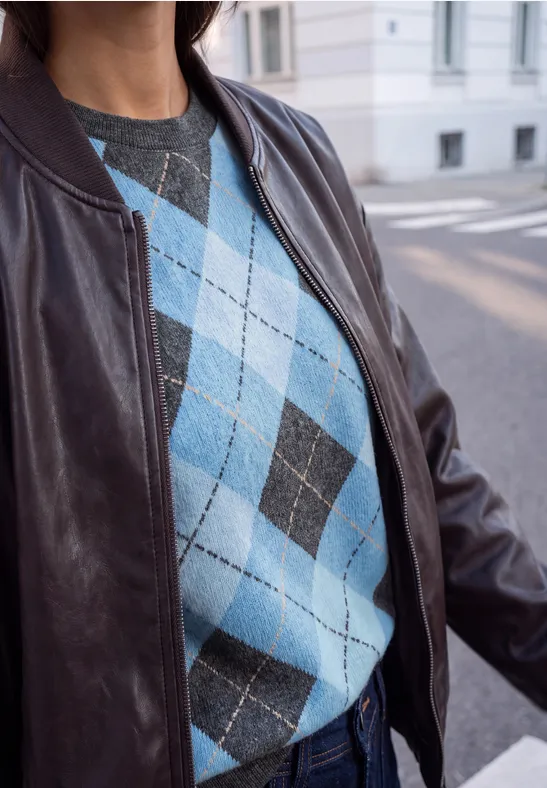Im Bild ist eine Person mit blau-grauem Rautenmuster-Pullover und brauner Lederjacke auf der Straße.