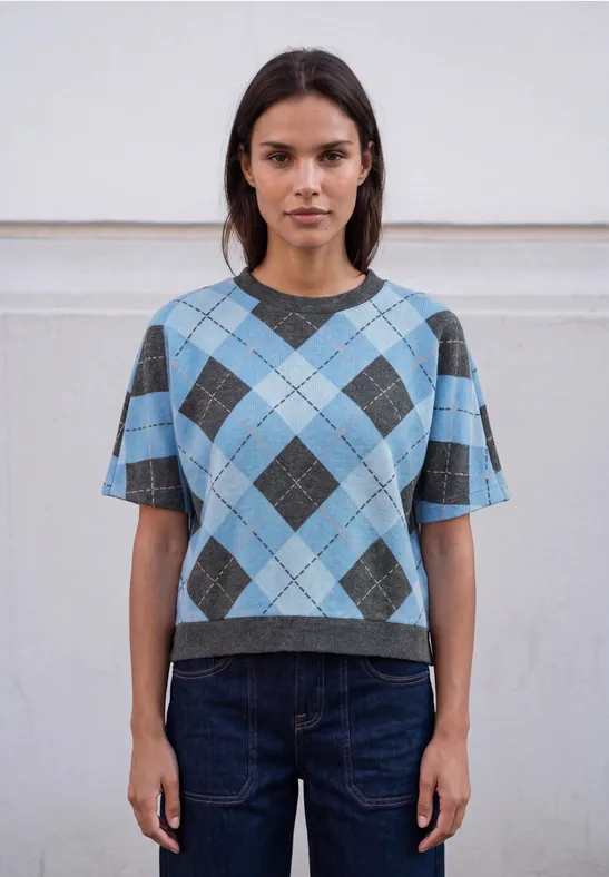 Frau trägt kurzes blau-graues Rauten-Pullover-Shirt und dunkle Jeans vor neutralem Hintergrund.