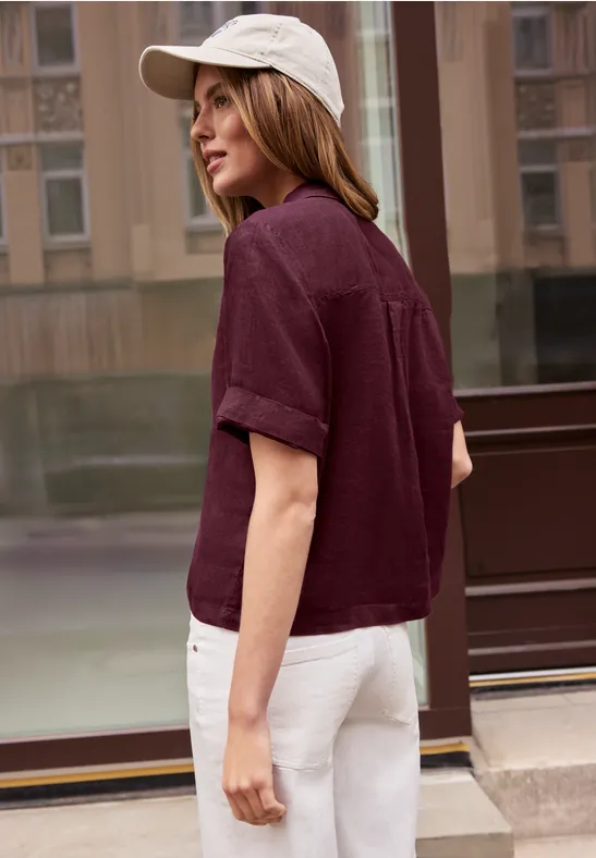 Une femme en chemisette courte bordeaux, pantalon blanc et casquette, debout de profil devant un immeuble vitré.