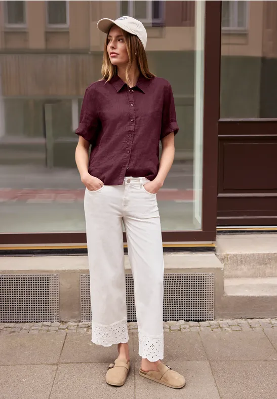 Femme en chemise bordeaux, pantalon blanc ample à dentelle et mocassins beiges, debout sur le trottoir.