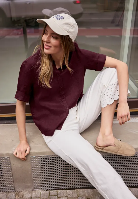 Une personne est assise de manière décontractée en pantalon blanc et t-shirt bordeaux devant une vitrine.
