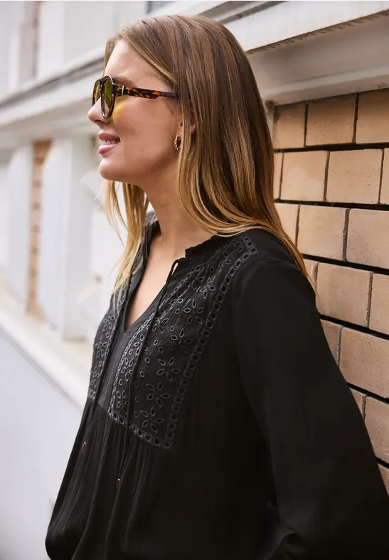 Frau mit langem Haar steht seitlich an einer Wand, trägt eine schwarze Bluse mit Spitzenbesatz.