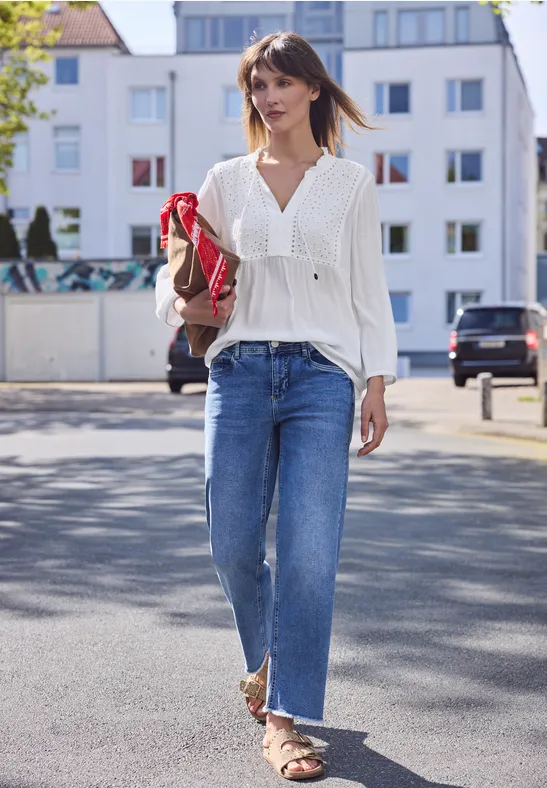 Een vrouw in een witte blouse, blauwe jeans en sandalen loopt met een tas over een zonnige straat.