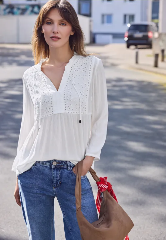 Vrouw in witte blouse en spijkerbroek met tas, staand in een zonnige straat.
