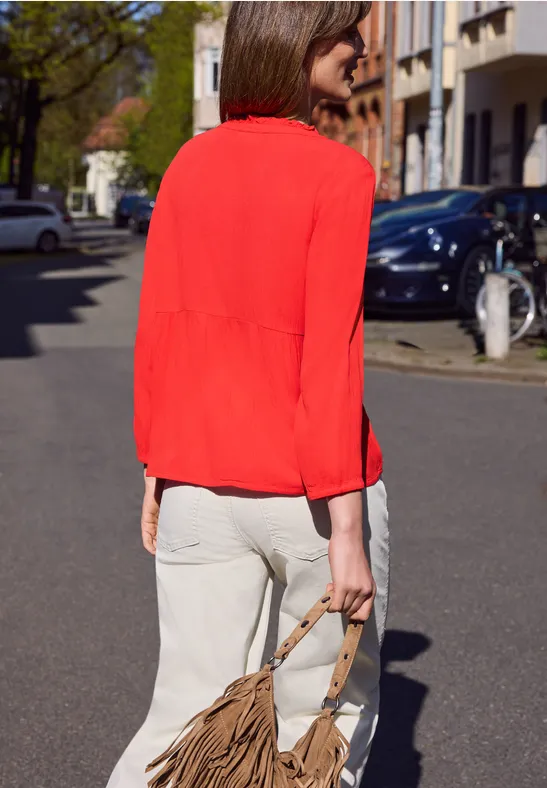 Een vrouw in een rode blouse en witte broek loopt op straat en draagt een bruine tas.