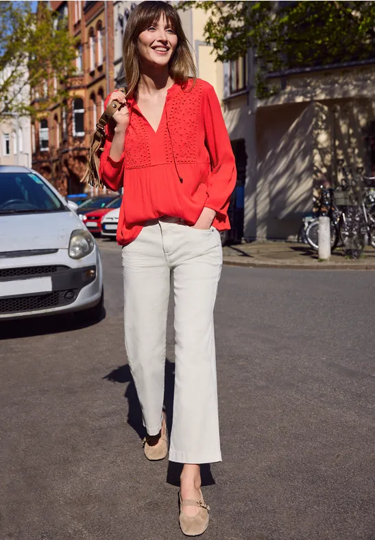 Een vrouw met een rode blouse en witte broek loopt over een zonnige straat.