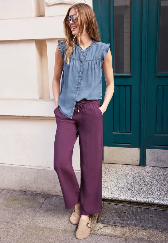 Vrouw in mouwloos blauw bloesje en wijde paarse broek staat ontspannen voor een voordeur.