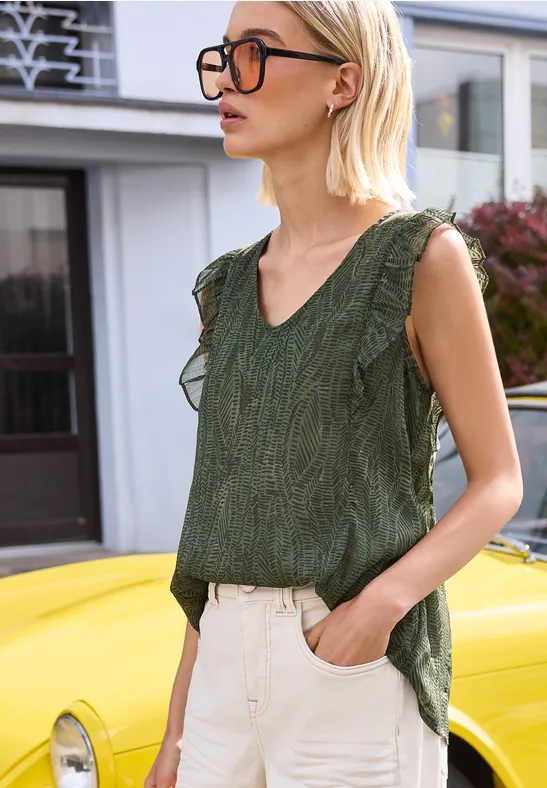 Une femme blonde avec lunettes et haut vert appuyée de façon décontractée sur une voiture jaune.