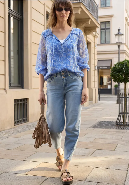 Vrouw in blauw gebloesd shirt, jeans en sandalen, met een franjes tas, lopend door een chique straat.