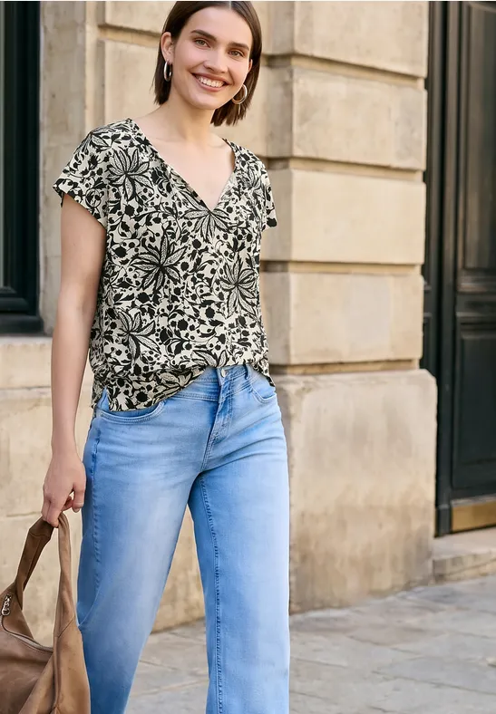 Vrouw in gemêleerd shirt, lichtblauwe spijkerbroek en tas, lopend langs een stenen muur.