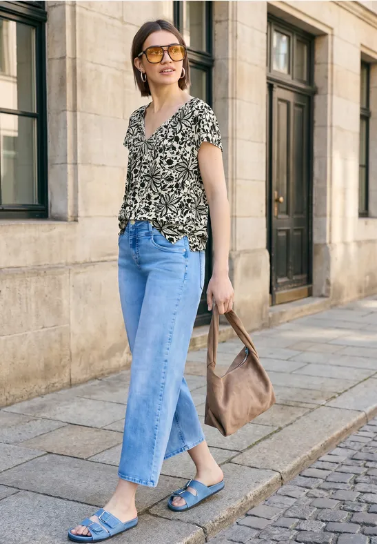 Vrouw in printshirt en jeans met sandalen en handtas loopt langs een gevel in de stad.