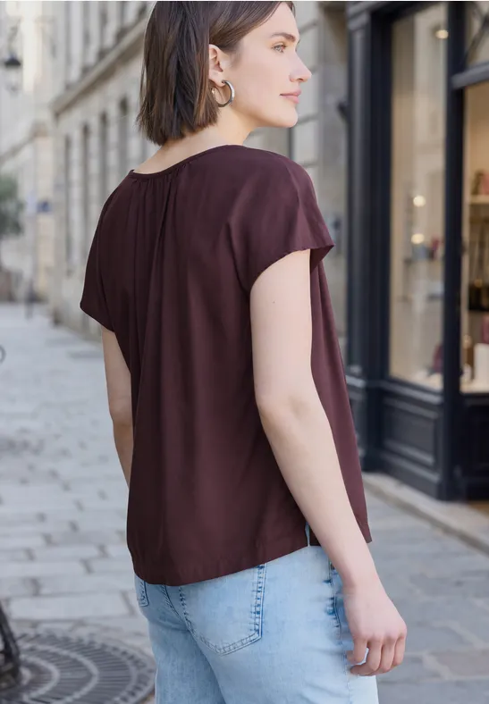 Frau in Burgunderfarbener Bluse und Jeans steht mit dem Rücken zur Kamera auf einer Kopfsteinpflasterstraße.