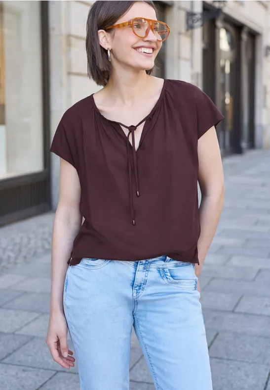 Frau in brauner Bluse mit V-Ausschnitt und hellblauen Jeans steht auf dem Bürgersteig.