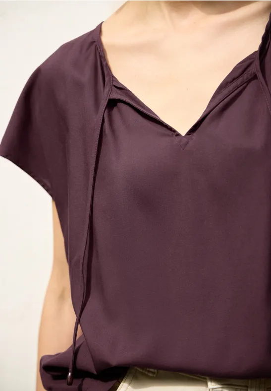 Lila Kurzarm-Bluse mit V-Ausschnitt und dekorativen Schlaufenbändern.