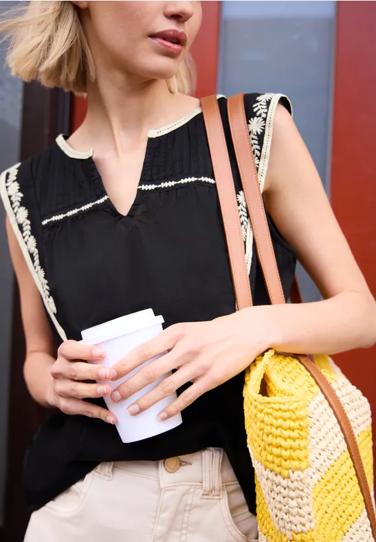 Eine Frau hält einen Pappkaffee und trägt eine gehäkelte gelbe Tasche, schwarzen Top und helle Hose.