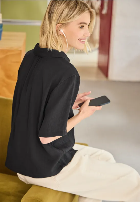 Frau sitzt auf Bank, hält Smartphone, trägt schwarzen Pullover und weiße Hose, Blick seitlich, indoor.