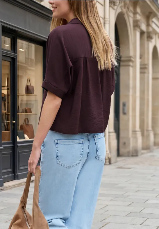 Eine Frau in hellblauen Jeans und dunkelrotem Oberteil trägt Tasche und spaziert an eleganten Gebäuden.