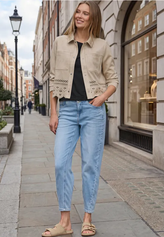 Frau in beiger Jacke und blauen Jeans geht lässig eine gepflasterte Einkaufsstraße entlang.