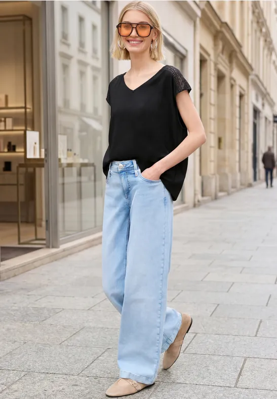 Femme en t-shirt noir et jean large se promène devant des vitrines élégantes.