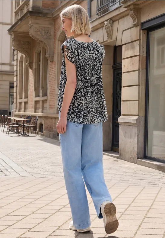 Frau mit schwarz-weiß gemustertem Shirt und hellblauen Jeans läuft auf gepflastertem Gehweg in urbaner Straße.