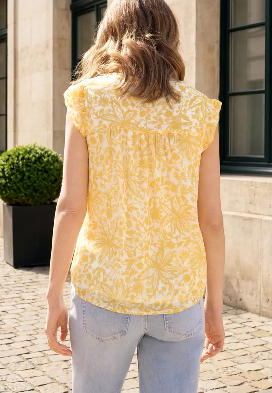 Eine Frau steht mit dem Rücken zur Kamera, trägt eine gelbe, gemusterte Bluse und Jeans vor einer Hauswand.