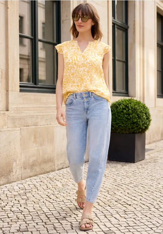 Frau in gelber Bluse, Jeans und Sandalen läuft auf gepflastertem Gehweg an Hausfassade mit Topfstrauch vorbei.