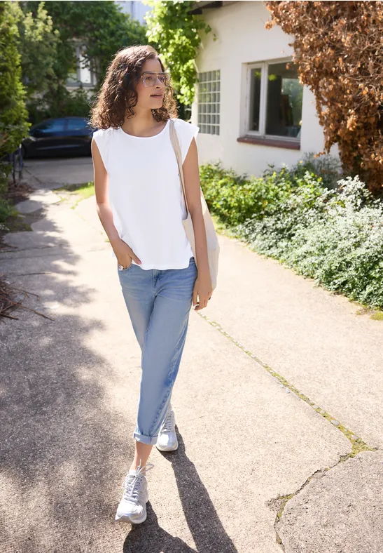Eine Frau in weißem Top, Jeans und Sneakers steht auf einem Gehweg vor einem Haus, umgeben von Grün.