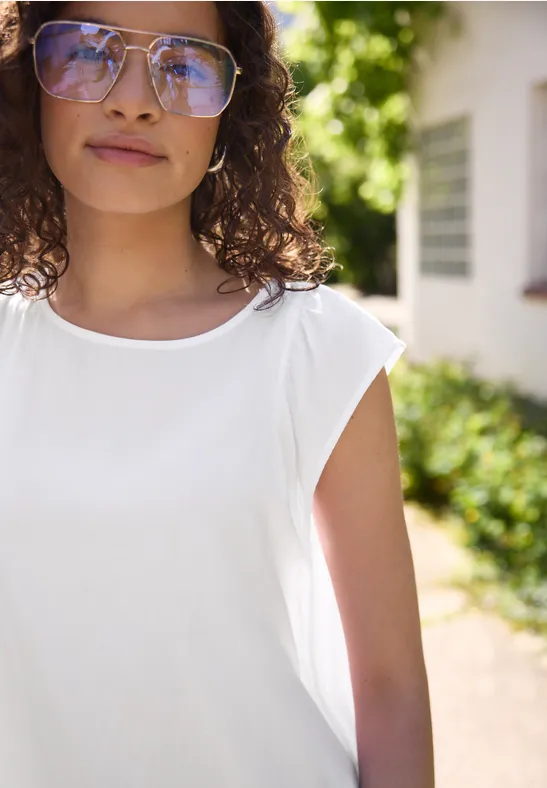 Eine Person mit lockigem Haar in weißem ärmellosen Oberteil im sonnigen Garten vor einem Haus.