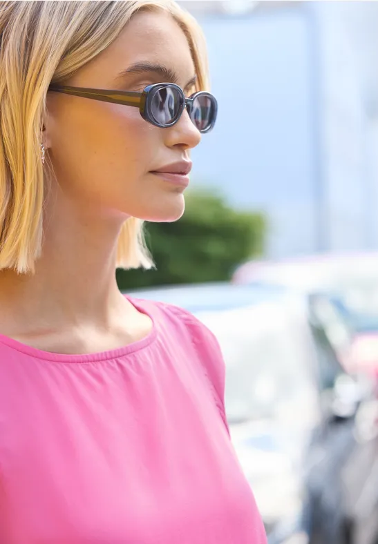Een vrouw met schouderlang blond haar draagt een roze top en staat buiten voor geparkeerde auto’s.