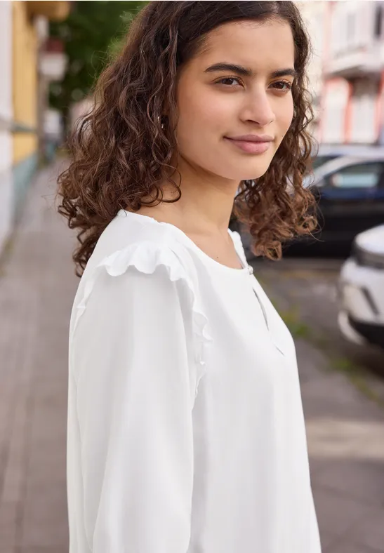 Eine Frau mit lockigem Haar trägt eine weiße Bluse und steht auf einem Bürgersteig vor geparkten Autos.