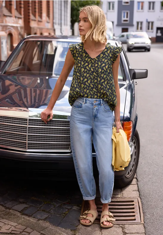 Een persoon met vervaagd gezicht leunt tegen een zwarte auto, draagt een bloemen top, lichte jeans en sandalen.