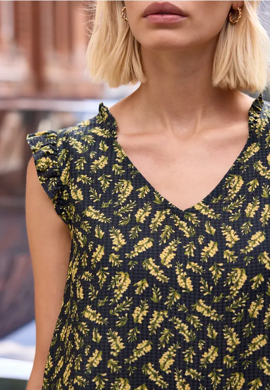 Een blonde vrouw in een zwarte, geel gebloemde jurk met V-hals en ruches aan de mouwen.