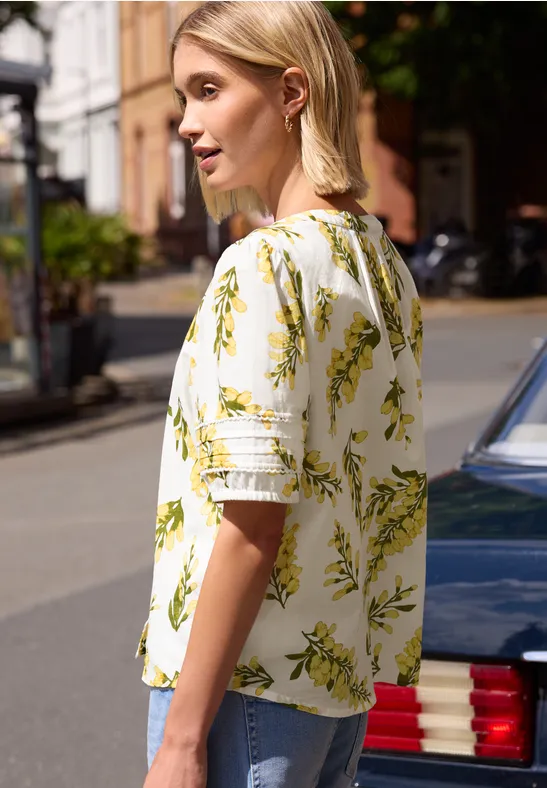 Zijaanzicht van persoon met wit bloesje met geel-groene bloemenprint naast auto op straat.