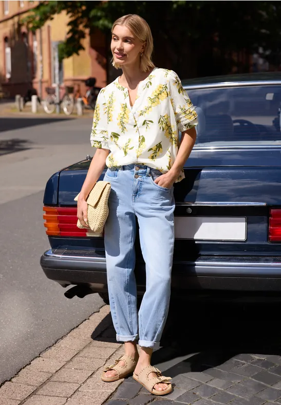 Vrouw in blouse met bladmotief en jeans leunt tegen een klassieke auto, draagt sandalen en houdt een clutch vast.