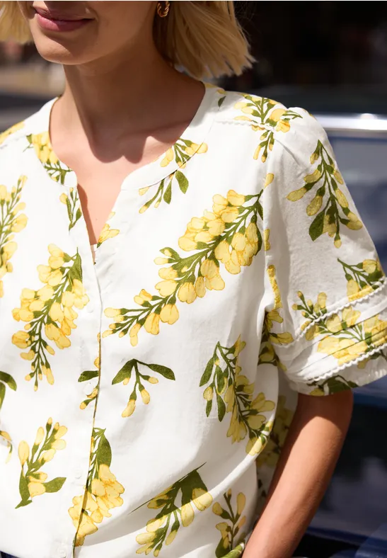 Een vrouw met blond haar draagt een witte blouse met gele bloemen en korte mouwen.