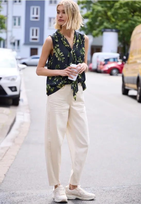 Frau steht in der Straße, trägt geblümtes Top, weite helle Hose und Slip-ons.