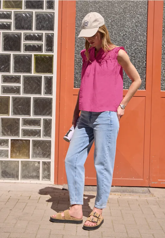 Frau in pinkem Top, Jeans und Sandalen vor einer roten Tür, mit weißer Kappe und Handy.