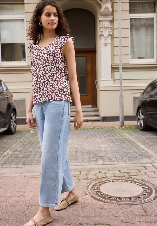 Eine Frau in geblümter Bluse und weiten Jeans steht auf einem gepflasterten Gehweg vor einem Gebäude.
