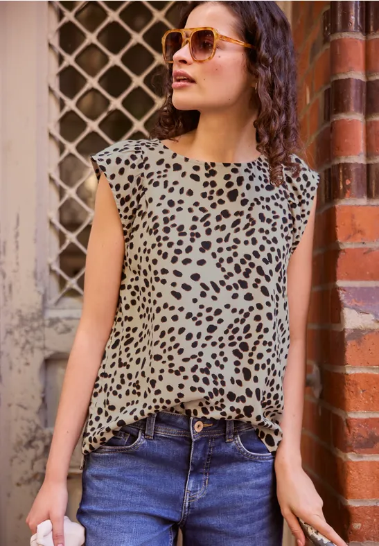 Vrouw leunt nonchalant tegen bakstenen muur, draagt jeans, mouwloos luipaardtop en zonnebril.