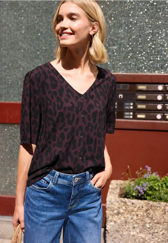 Frau in schwarz-violettem Leoparden-V-Shirt und Jeans steht vor einer Hausfassade mit Briefkästen.
